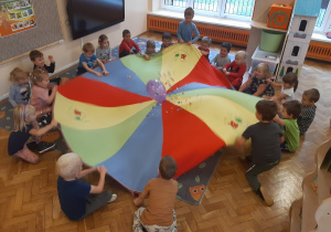 Dzieci ustawione w koło trzymają rękoma chustę animacyjną. Podrzucają umieszczone na chuście pompony i balon.